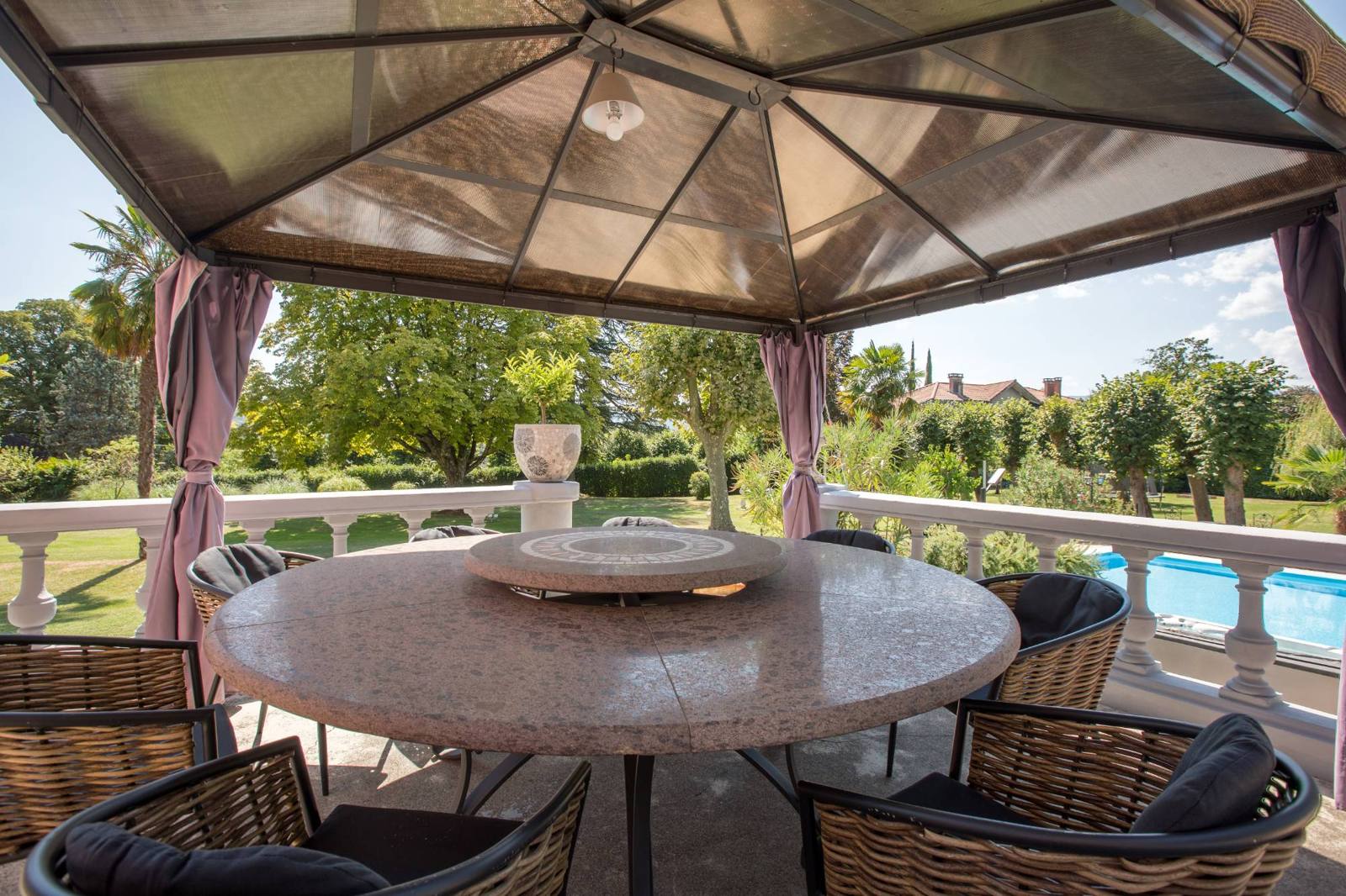 A round stone table and wicker chairs under a curtained pavilion, overlooking the gardens and pool