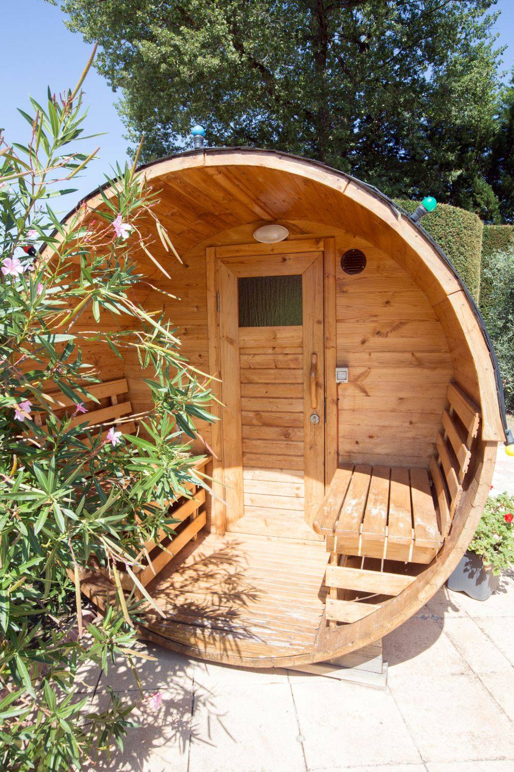 A wooden barrel sauna with benches, framed by a flowering oleander