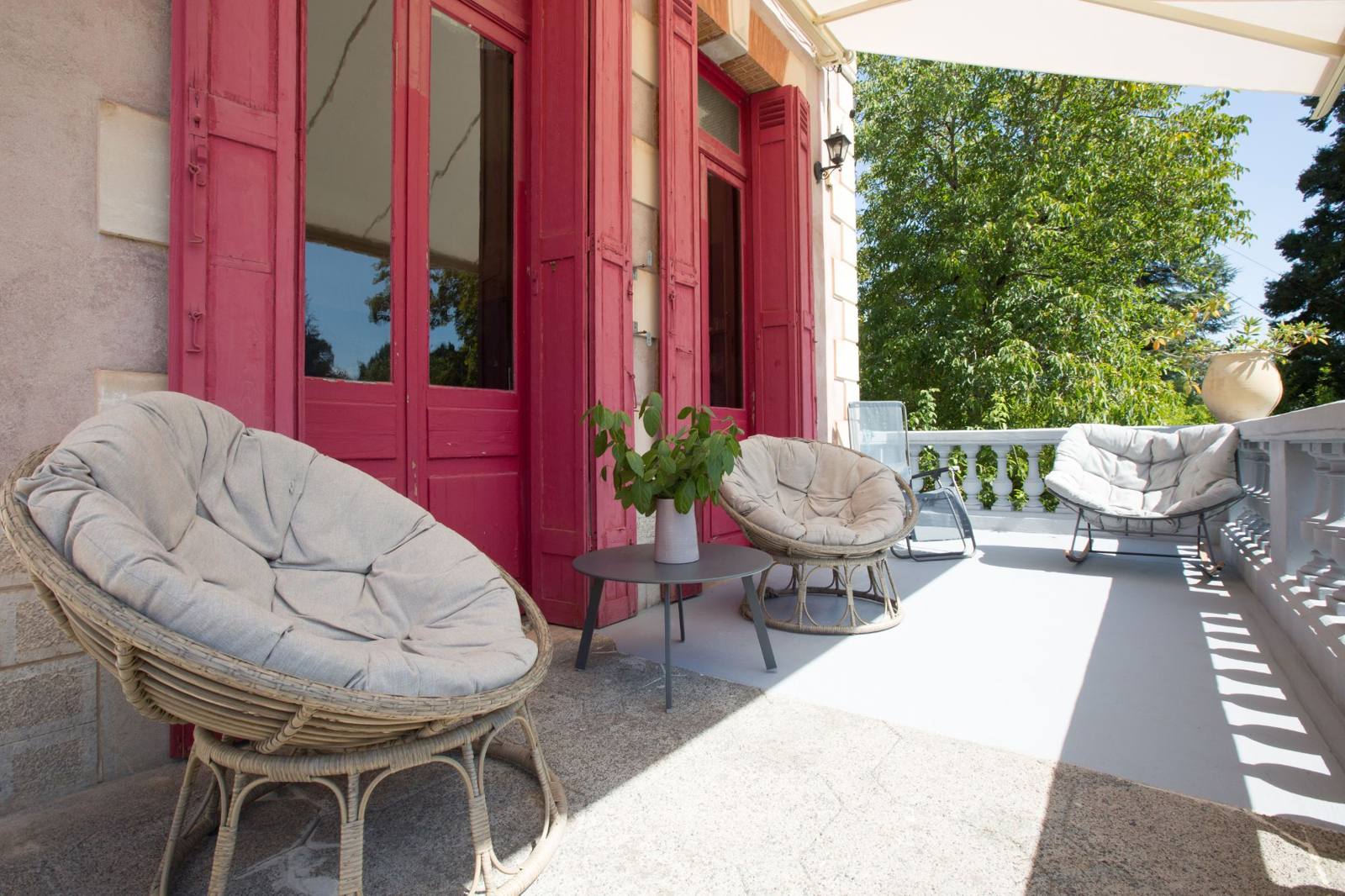 Wicker armchairs on a sunny terrace beside tall raspberry-red shutters and a stone balustrade