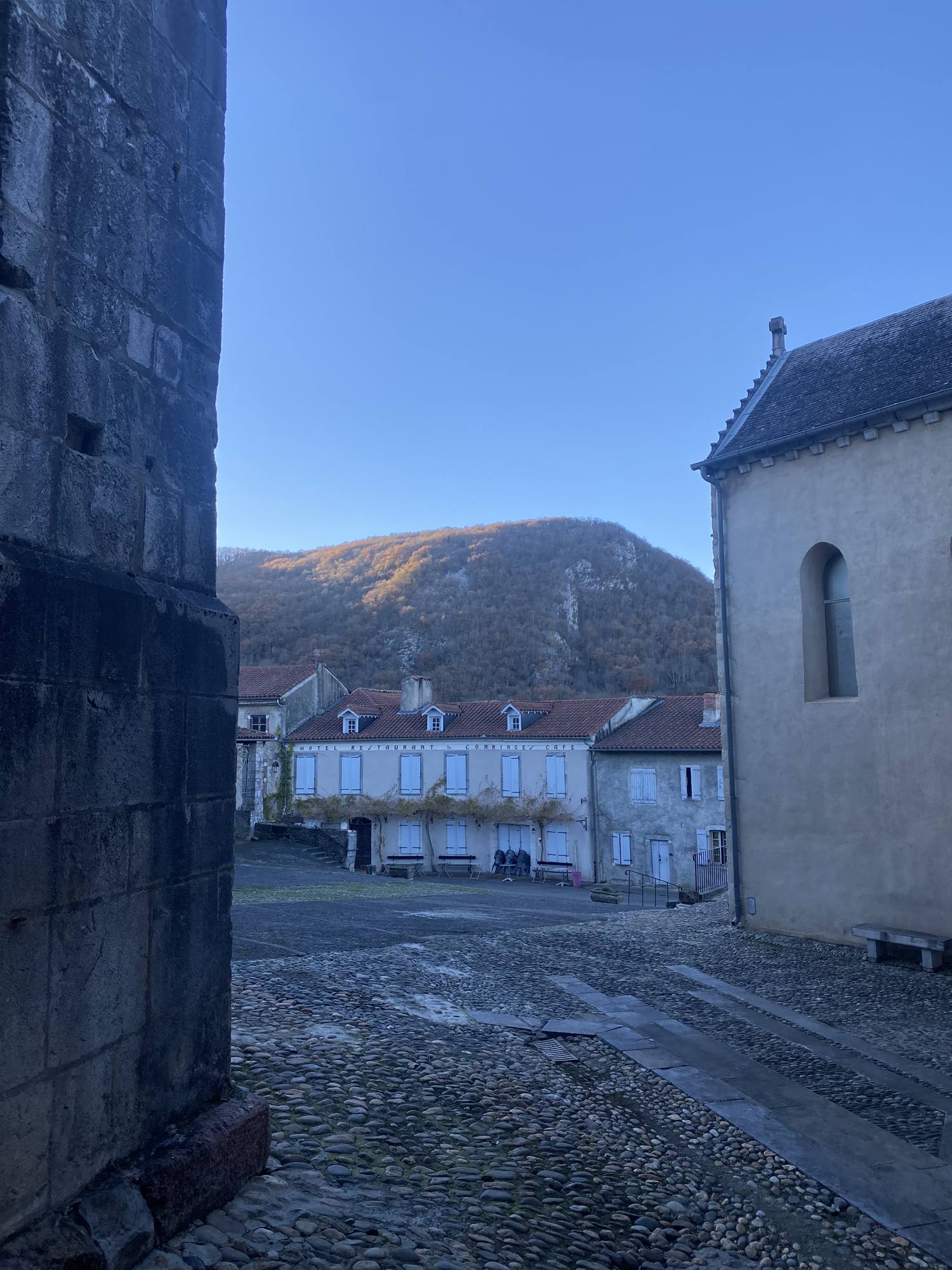 A cobbled village square beside an old stone church, with a wooded hill behind