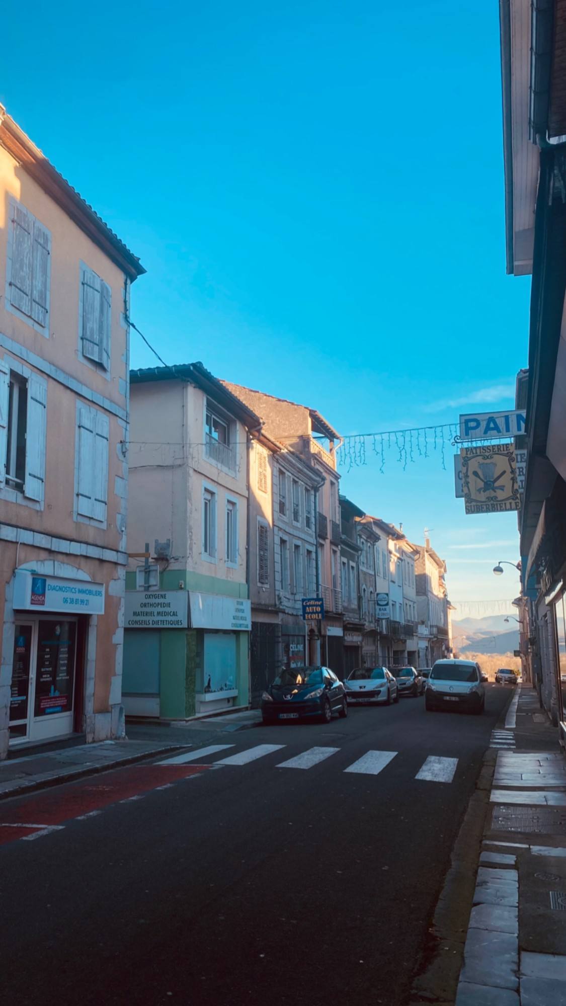 A street of tall shuttered townhouses with a bakery sign, leading towards the Pyrenees in winter sunshine